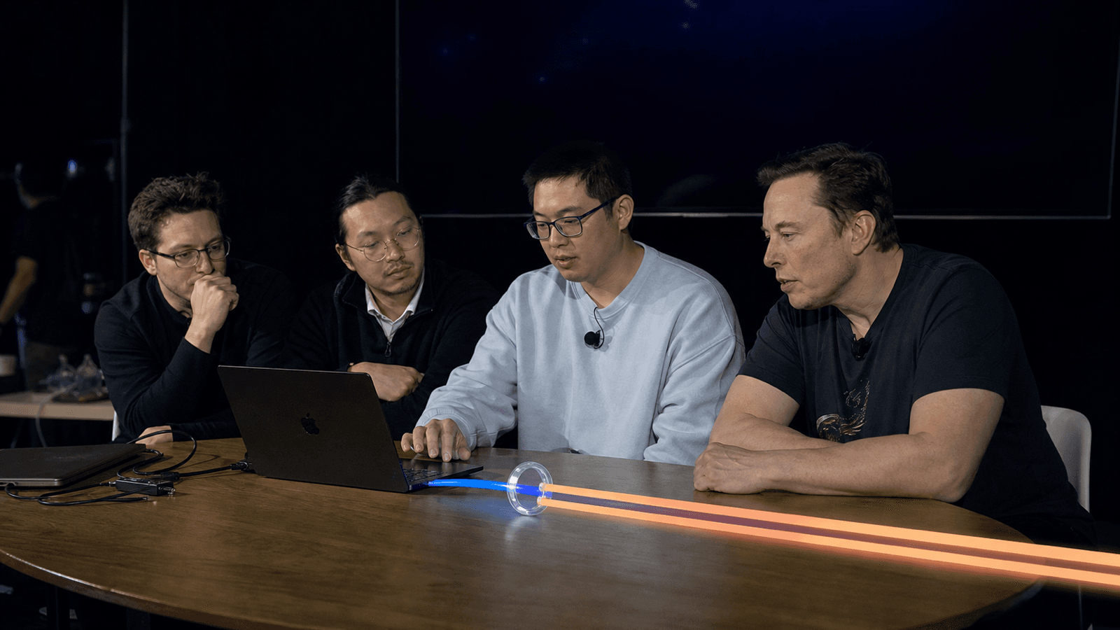 Four men sit along one side of a table in a darkened studio, watching a laptop one of them is typing on. A narrow blue line of light runs out of the laptop into a small glass lens on the table, where it re-emerges as two far broader orange beams that widen away across the surface. The man at the right of the group is depicted as Elon Musk.