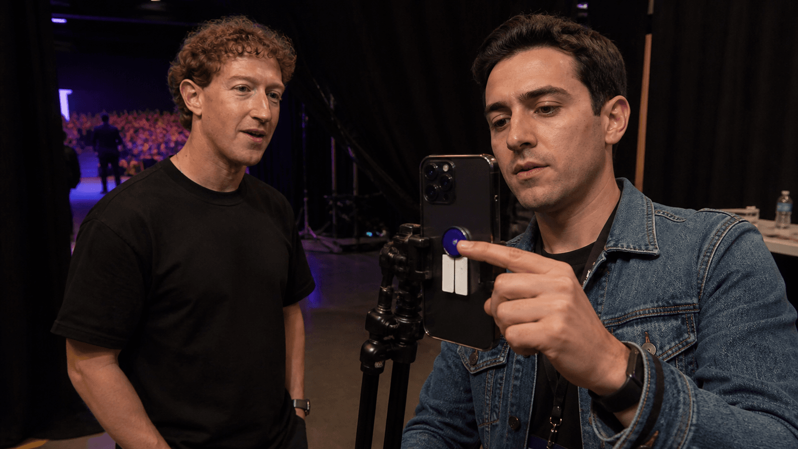 Two men stand backstage behind a dark curtain with a lit auditorium visible through the gap beyond. The man at left, depicted as Mark Zuckerberg, watches as a younger man in a denim jacket reaches past a phone mounted on a tripod rig and points at a small blue control on its side.