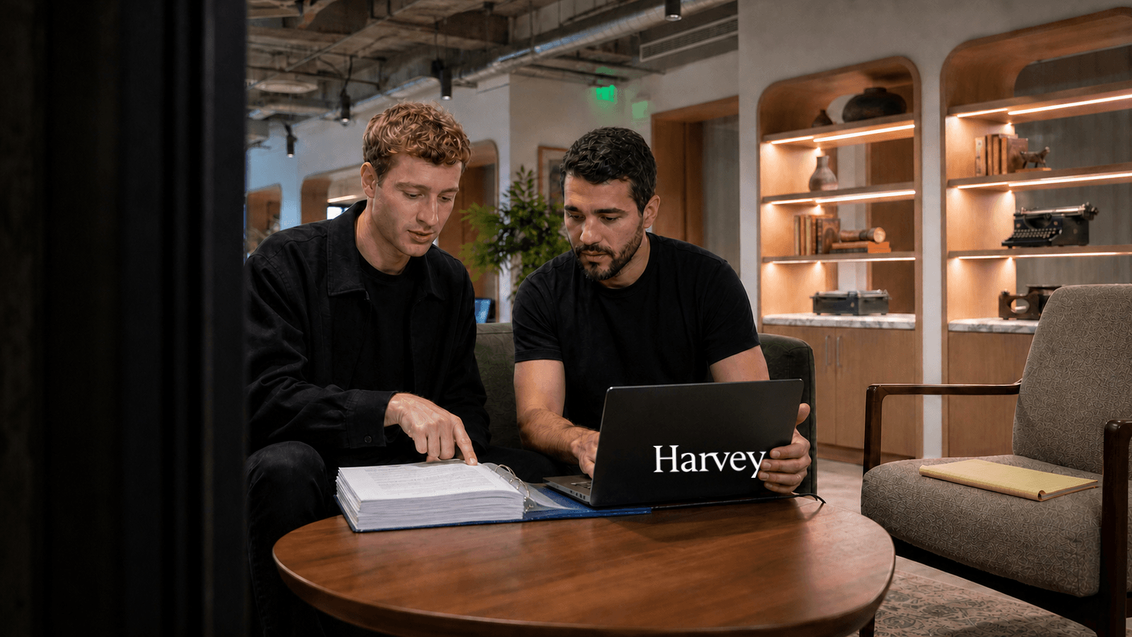Two men in black sit together on a couch in a softly lit office lounge. One points at a page in a thick ring binder of printed documents open on the low table; the other works on a laptop whose lid carries the Harvey wordmark. Behind them, lit shelving holds a vintage typewriter and other antique office machines.