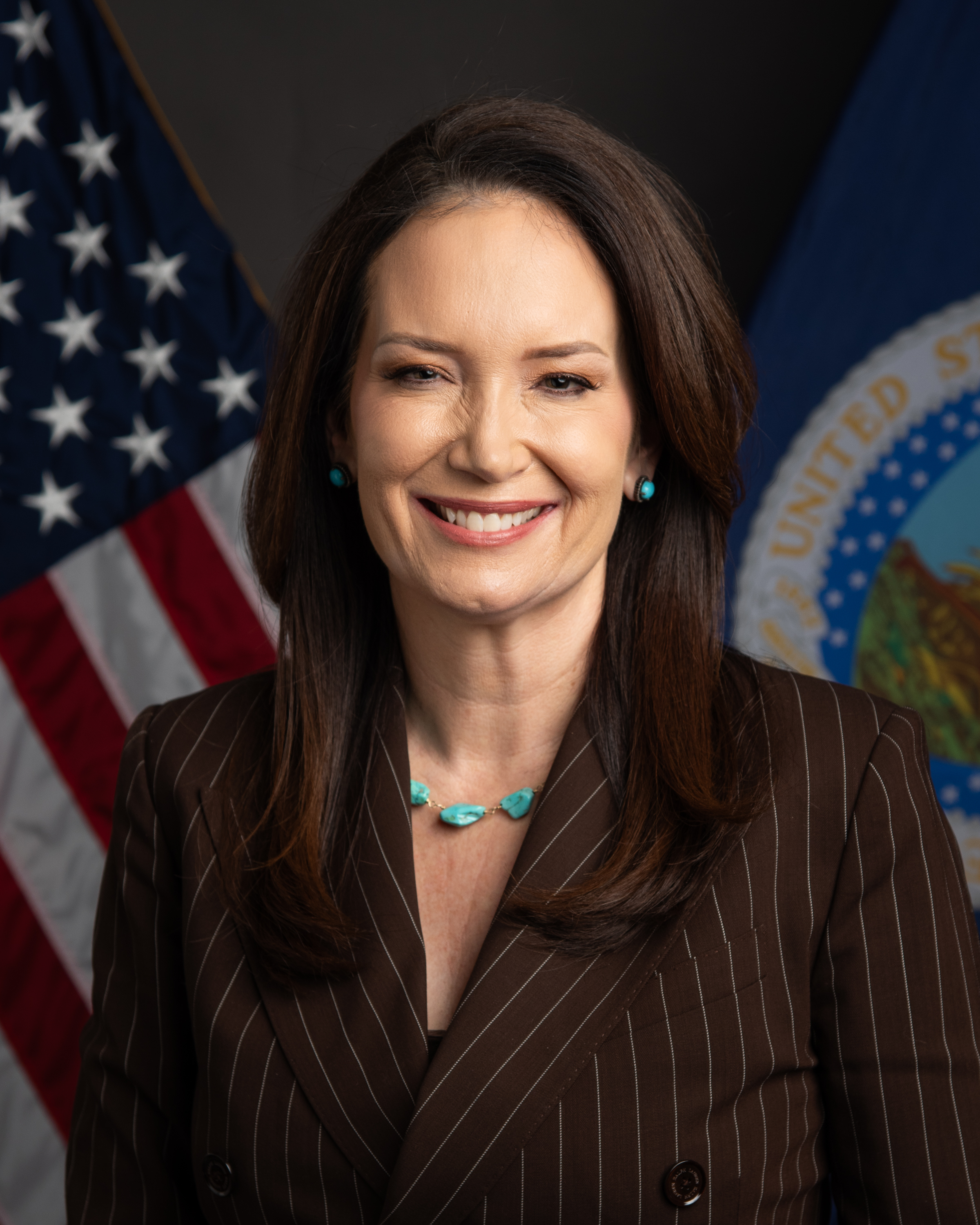 Brooke Rollins, Secretary of Agriculture, smiling in a brown pinstriped jacket and a turquoise necklace, in front of an American flag and a department flag.